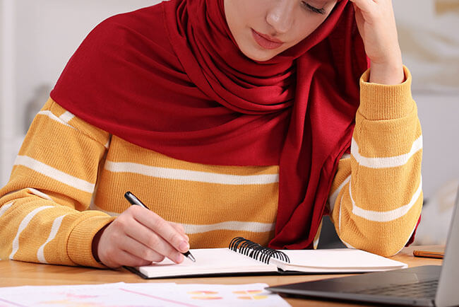 Woman in a red hijab thoughtfully working in front of a laptop.