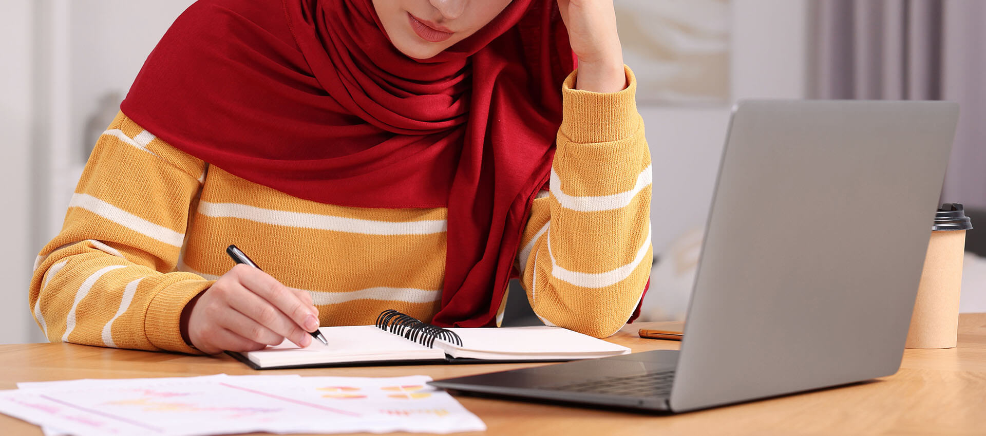 Woman in a red hijab thoughtfully working in front of a laptop.