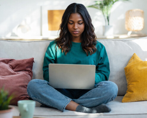 A BIPOC woman sits cross legged on a couch looking at her laptop